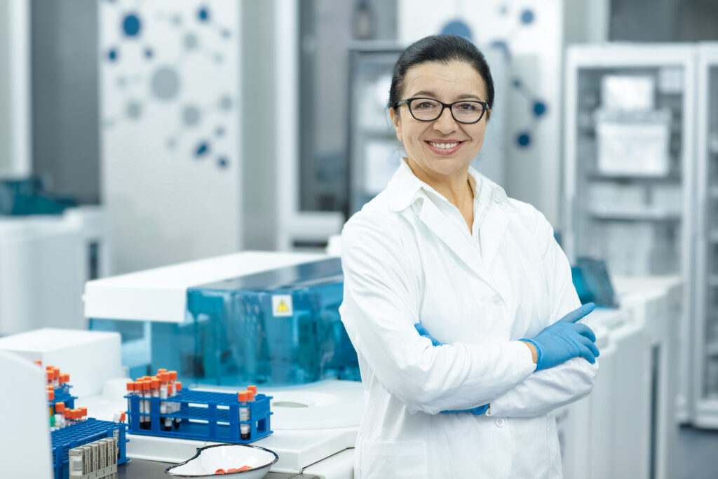 Senior specialist. Cheerful elderly female scientist smiling happily with her arms crossed posing at her lab colleagues on the background teamwork profession professor guidance people job copyspace